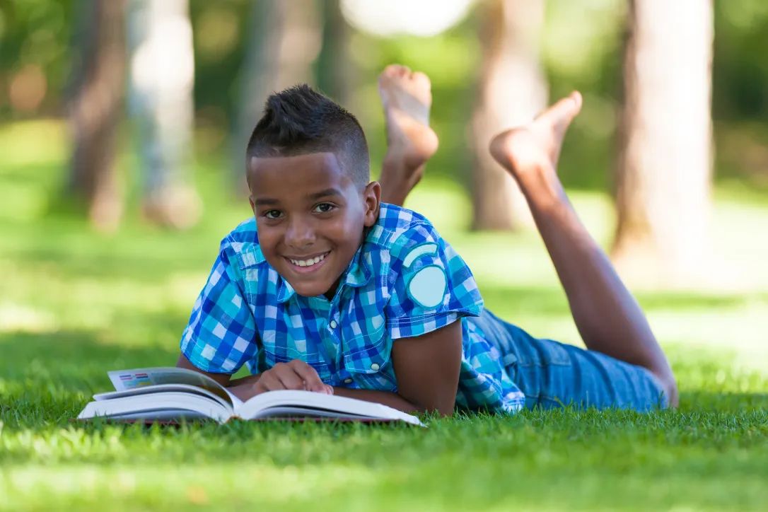 A smiling boy lying on the grass reading a book
